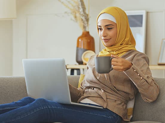 Woman in a yellow hijab sits on a couch, holding a mug and using a laptop, with a vase and framed picture in the background.