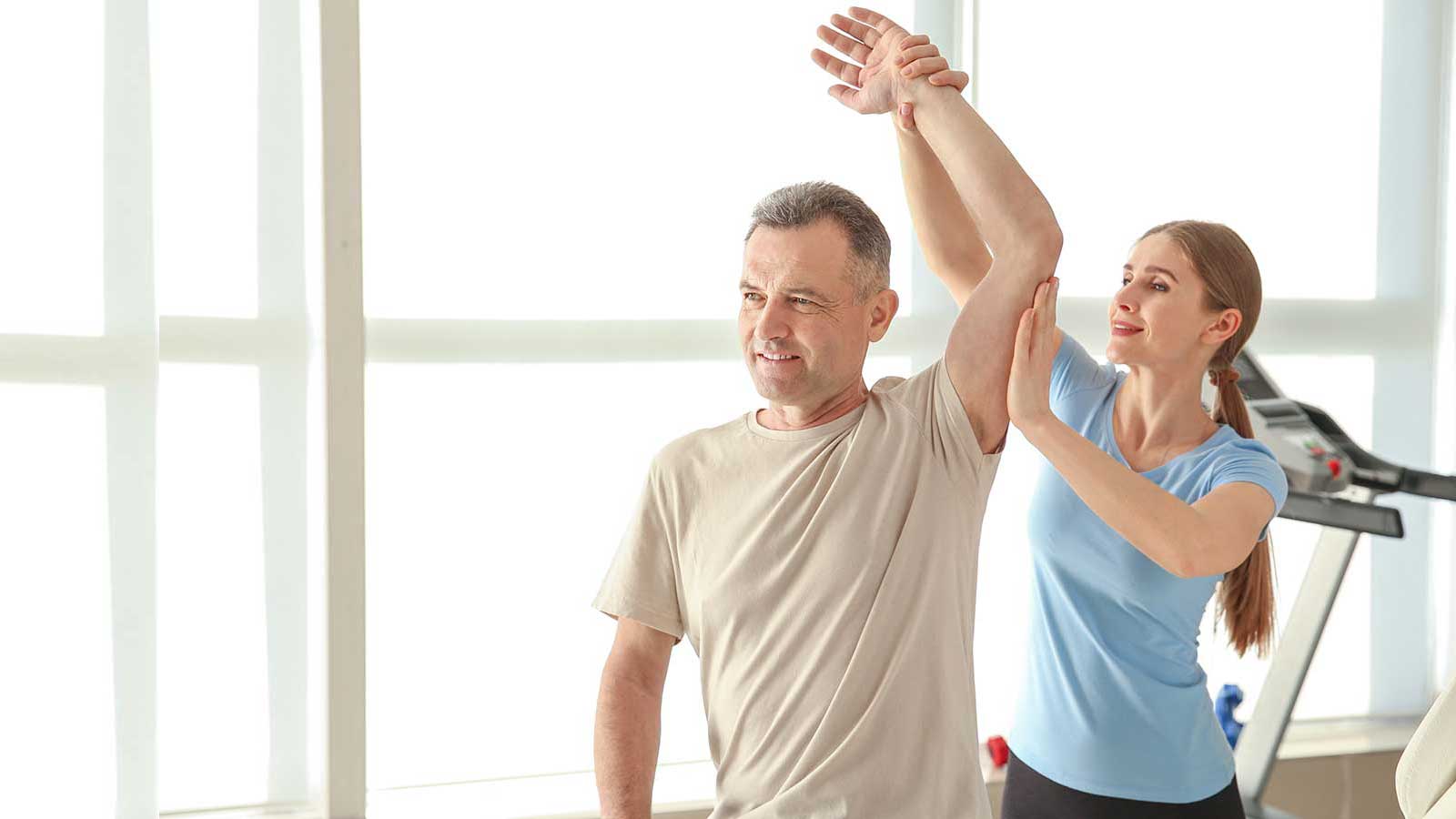 A man in a beige shirt stretches his arm overhead with assistance from a woman in a blue shirt in a bright gym.