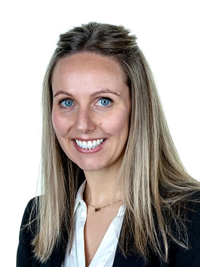 Smiling woman with long blonde hair wearing a black blazer and white blouse against a plain background.
