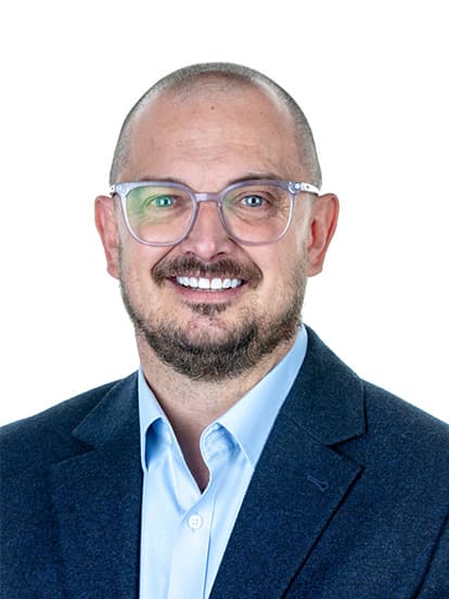 A smiling man with glasses, bald head, and beard wearing a blue shirt and dark blazer