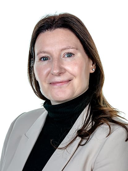 A woman with long brown hair wearing a beige blazer and black turtleneck, smiling at the camera against a white background.