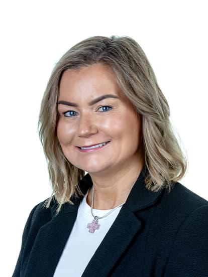 A woman with shoulder-length blonde hair, wearing a black blazer, white top, and a cross necklace, smiling