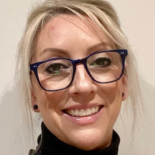 Smiling person with blue glasses and blonde hair, wearing a black turtleneck, stands against a neutral background.