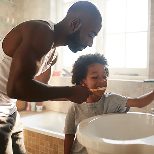 A man helps a child brush their teeth in a sunlit bathroom, standing by the sink with a window in the background.