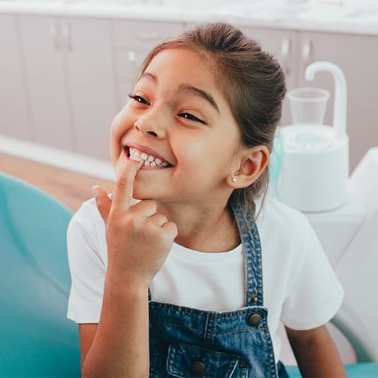 Smiling girl in a dental office, pointing at her teeth. She wears a white shirt and denim overalls, sitting on a blue dental chair.