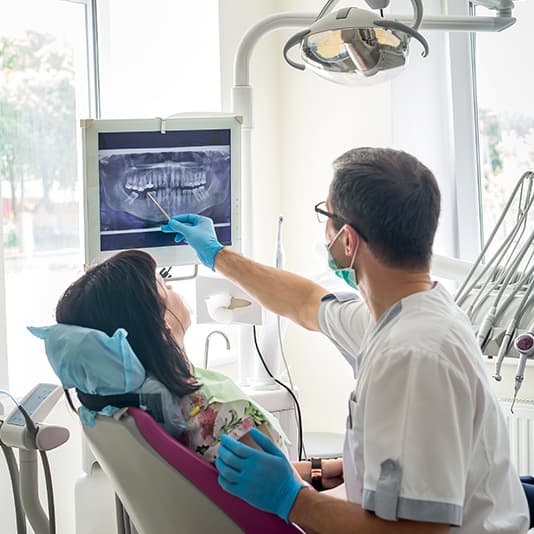 Dentist showing female patient a dental X-ray on a screen, pointing with a tool, in a brightly lit dental office.