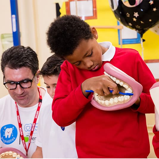 Child in red jumper brushing a large dental model while an adult supervises, in an educational setting with bright walls.