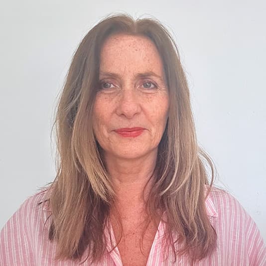 A woman with long brown hair and a pink striped shirt, standing against a plain white background, smiles softly at the camera.
