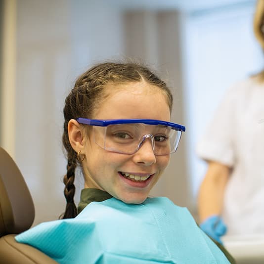 Smiling girl with braids wearing protective glasses and a dental bib, sitting in a dental chair with a blurred professional in the background.