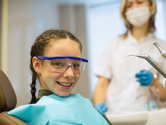 A smiling girl in a dental chair wearing protective eyewear and a bib, with a masked dentist in the background holding dental tools.