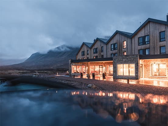 A modern wooden hotel with warm lights reflecting on wet ground, set against a misty mountain backdrop under a cloudy sky.