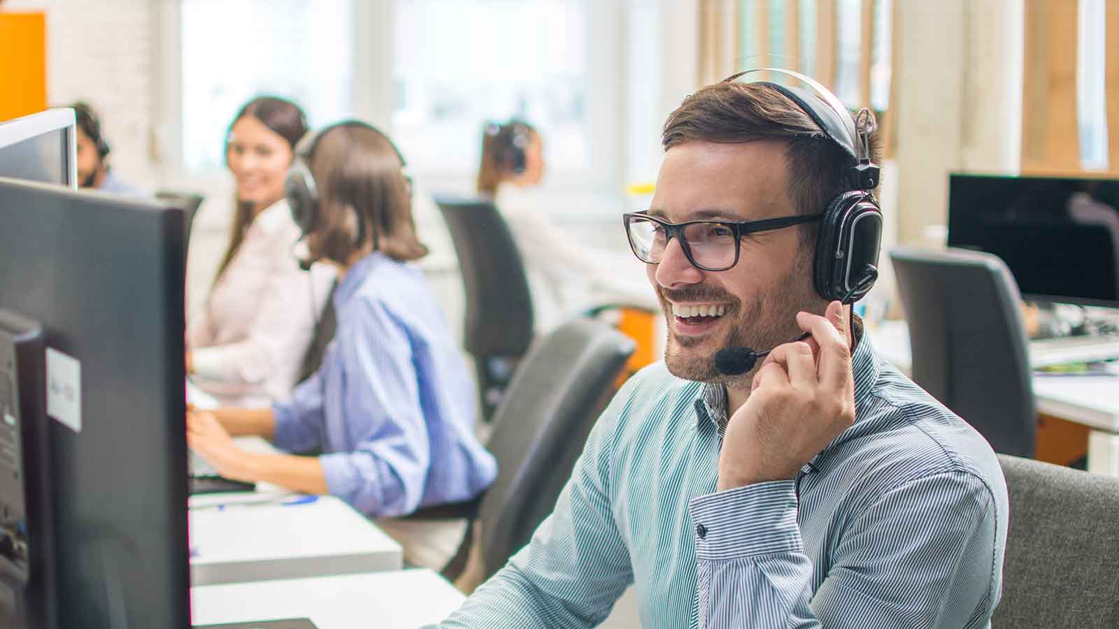 Man wearing glasses and headset smiles while working at a computer in a busy office, with colleagues in the background.