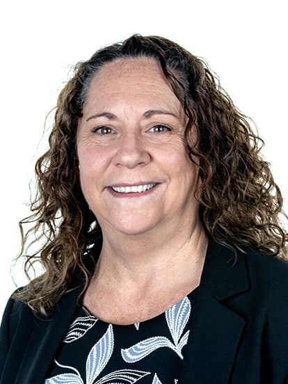 A woman with curly brown hair smiles warmly, wearing a black blazer and a patterned blouse