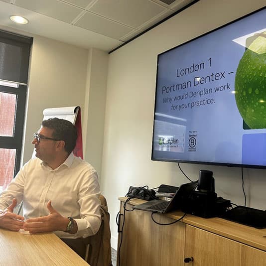 Man in a meeting room presenting with a slide on the screen titled "London 1 Portman Dentex." A flip chart is visible in the background.