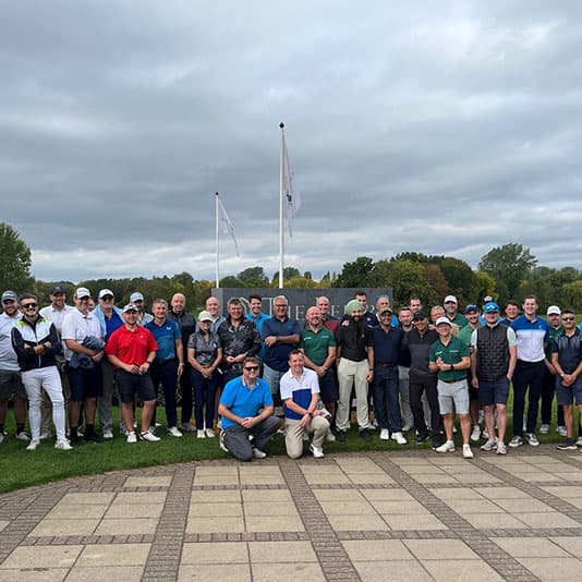 A large group of people posing on a golf course, with flags in the background and cloudy skies above.