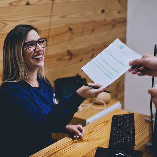 A smiling woman at a wooden desk hands a document to someone. She is wearing glasses and holding a pen, with a computer keyboard nearby.