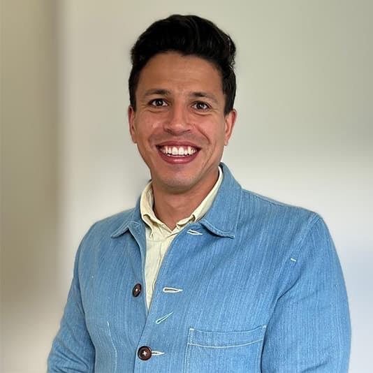 Smiling person with short dark hair wearing a blue jacket poses against a plain background.