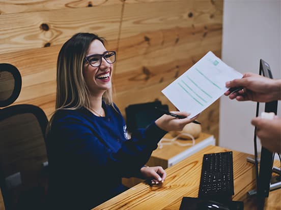 A smiling woman in glasses sitting at a wooden desk hands a document to someone. She is wearing a blue shirt and seated in an office with wood-paneled walls.