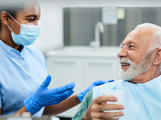 A dental professional wearing a mask and gloves talks to an elderly man holding a cup, both smiling in a dental clinic setting.