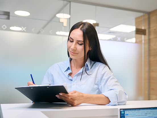 Woman in a light blue shirt writing on a clipboard at a desk, in a modern room with glass walls and lights in the background.